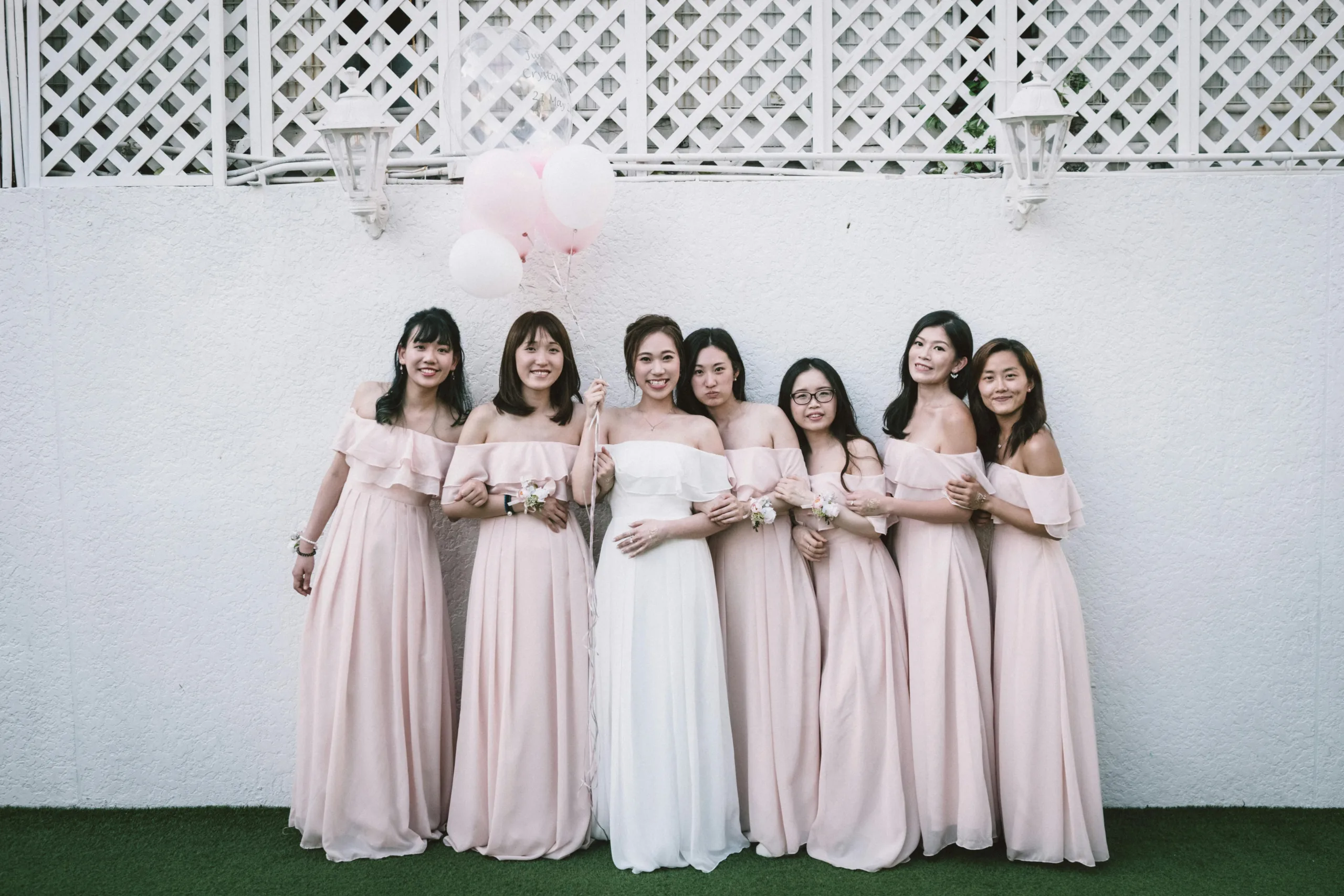 Cheerful young Asian bride with balloons and bridesmaids in butifull dresses standing on grass near white wall and looking at camera