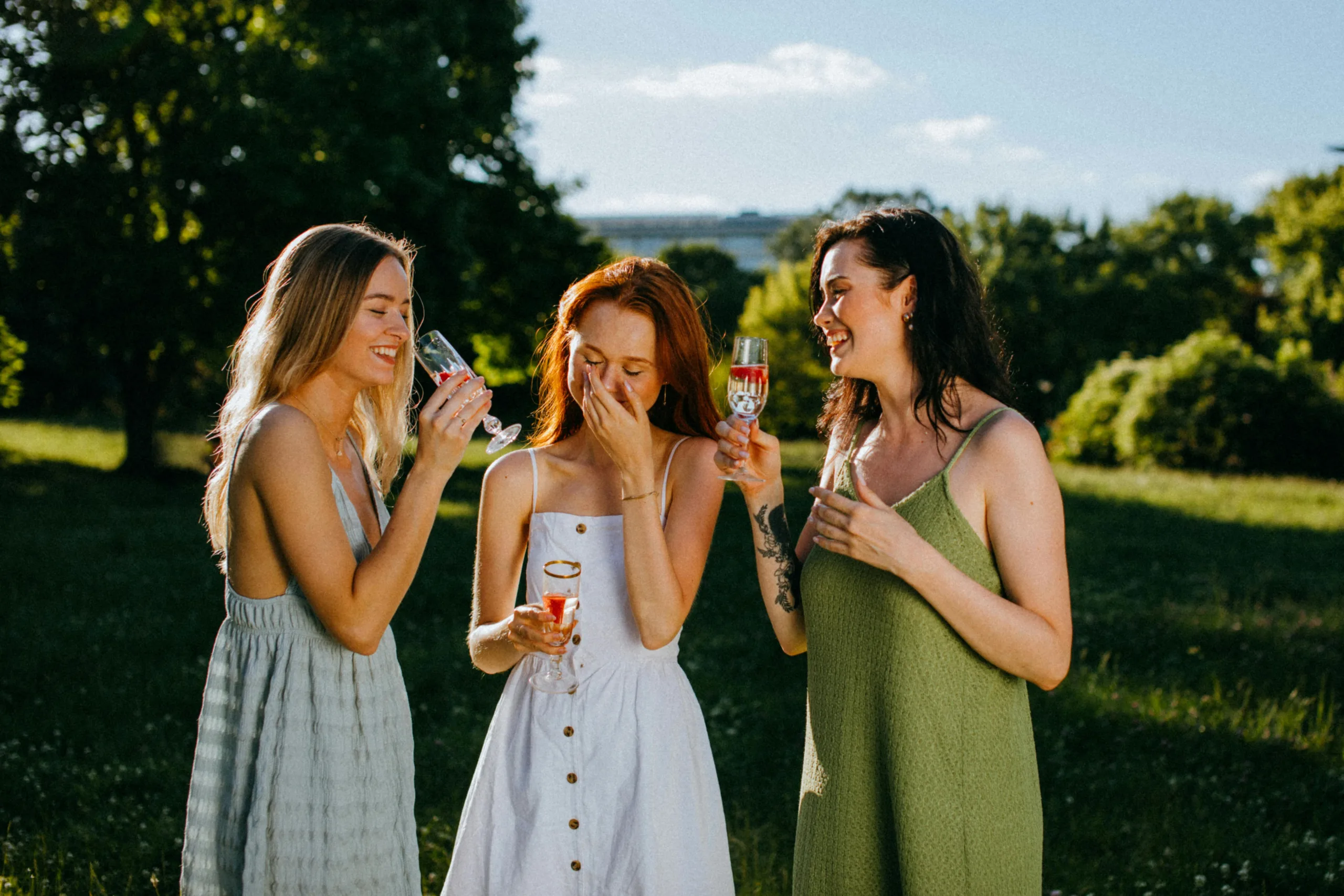 Three women enjoying a sunny day with drinks, celebrating friendship outdoors.